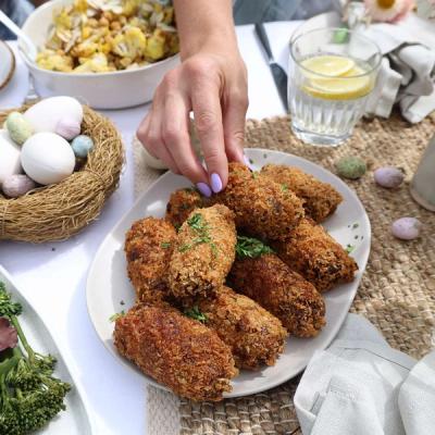 A hand with lavender nail polish picks a golden-brown croquette from a plate, surrounded by a salad bowl, a nest of white and pastel Easter eggs, and a glass of lemon water.