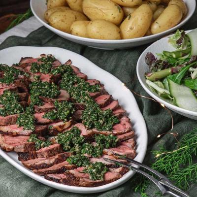 A platter of sliced steak topped with chimichurri sauce, served with bowls of potatoes and salad. A platter of sliced steak topped with chimichurri sauce, served with bowls of potatoes and salad.