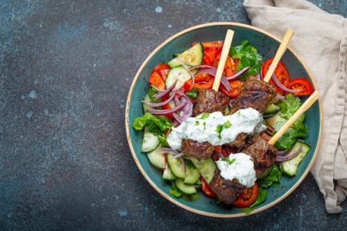 a bowl of food with meat skewers and vegetables on a table . a bowl of food with meat skewers and vegetables on a table .