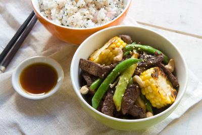 Beef stir-fry with corn, green beans, cashews, and sesame seeds, served with white rice, chopsticks, and soy sauce.