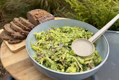 A bowl of green salad with a serving spoon, next to a sliced loaf of dark bread on a wooden board outdoors. A bowl of green salad with a serving spoon, next to a sliced loaf of dark bread on a wooden board outdoors.