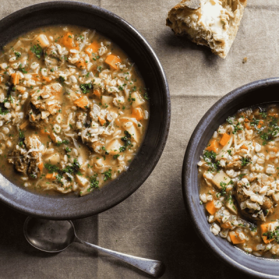 Two bowls of hearty meat and barley soup with vegetables and parsley, next to a piece of buttered bread. Two bowls of hearty meat and barley soup with vegetables and parsley, next to a piece of buttered bread.