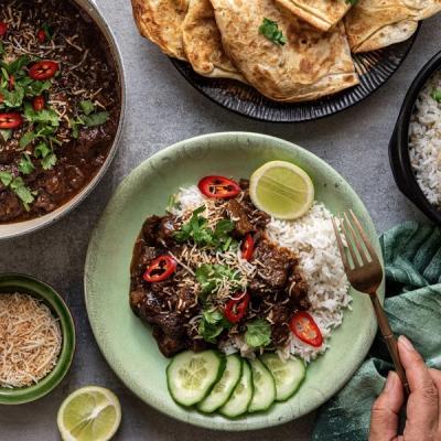 A plate of beef rendang with rice, cucumber, lime, chilies, cilantro, and toasted coconut, with a pot of rendang, flatbread, and a rice bowl in the background. A plate of beef rendang with rice, cucumber, lime, chilies, cilantro, and toasted coconut, with a pot of rendang, flatbread, and a rice bowl in the background.