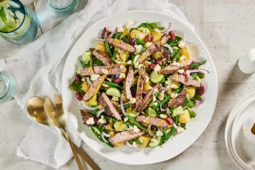 A large salad on a white platter featuring sliced lamb, potatoes, green beans, red beans, cucumber, red onion, and crumbled feta cheese, drizzled with dressing. A large salad on a white platter featuring sliced lamb, potatoes, green beans, red beans, cucumber, red onion, and crumbled feta cheese, drizzled with dressing.