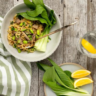 A bowl of ramen noodles with lamb, edamame, and bok choy on a rustic wooden table, next to a glass of lemon water and lemon slices.