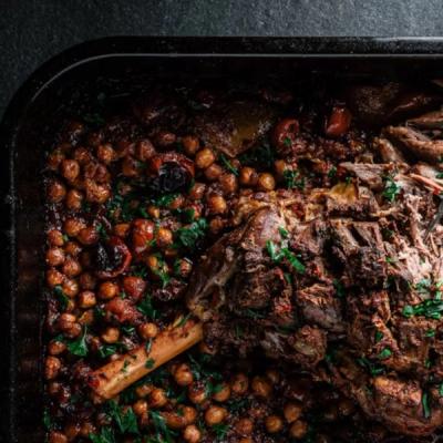 A roasted lamb shank with chickpeas, tomatoes, and parsley in a dark baking pan.