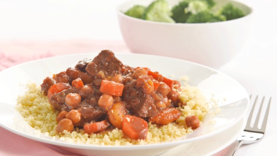 A plate of couscous topped with a meat, chickpea, and carrot stew, with a bowl of broccoli in the background.