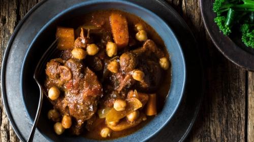 A blue bowl of rustic stew with meat, chickpeas, and carrots, on a wooden table with a side of broccoli.