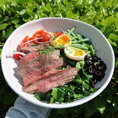 a person is holding a bowl of salad with steak , eggs , green beans , olives and tomatoes . a person is holding a bowl of salad with steak , eggs , green beans , olives and tomatoes .