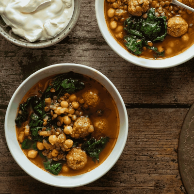 Two bowls of meatball, lentil, chickpea, and spinach soup, served with a side of yogurt and lemon wedges on a wooden table.