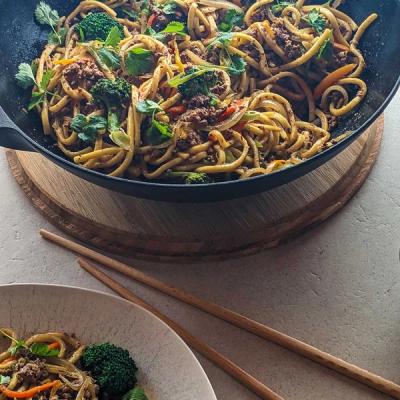 a wok filled with noodles. meat and vegetables next to a plate of noodles and chopsticks. a wok filled with noodles. meat and vegetables next to a plate of noodles and chopsticks.