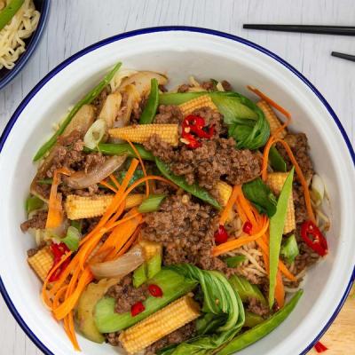 A white bowl with a blue rim filled with beef mince stir-fry, noodles, baby corn, carrots, green vegetables, and red chili garnish.
