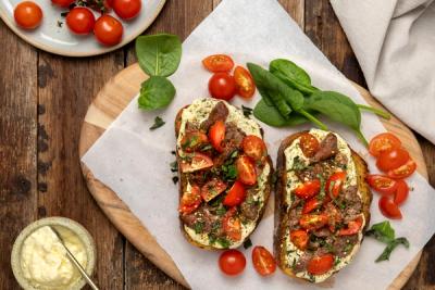 Two toasts topped with lamb, tomatoes, and creamy herb sauce, with fresh spinach and cherry tomatoes on a wooden board.