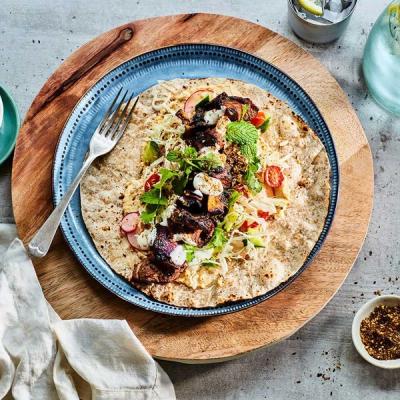A flatbread wrap filled with dark grilled meat or vegetables, hummus, shredded cabbage, radishes, and fresh herbs, served on a blue plate. A flatbread wrap filled with dark grilled meat or vegetables, hummus, shredded cabbage, radishes, and fresh herbs, served on a blue plate.