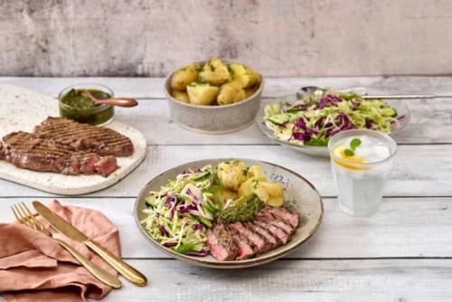 Sliced grilled steak, roasted potatoes, and a colorful cabbage salad with green sauce and lemon water.