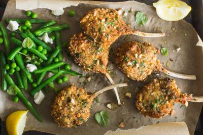 Five breaded lamb chops with green beans, peas, and a lemon wedge on parchment paper. Five breaded lamb chops with green beans, peas, and a lemon wedge on parchment paper.