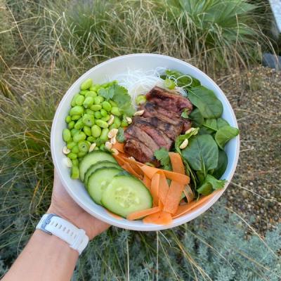 A hand holds a bowl of food containing sliced grilled steak, edamame, cucumber, carrots, spinach, rice vermicelli noodles, and peanuts.