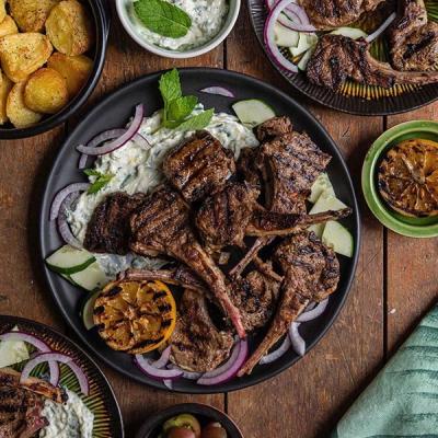 Grilled lamb chops with tzatziki, red onion, cucumber, and grilled lemon on a black plate, amidst other dishes on a wooden table. Grilled lamb chops with tzatziki, red onion, cucumber, and grilled lemon on a black plate, amidst other dishes on a wooden table.