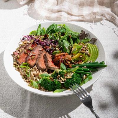 A vibrant bowl meal with sliced steak, brown rice, avocado, broccoli, green beans, and mixed greens, topped with seeds.