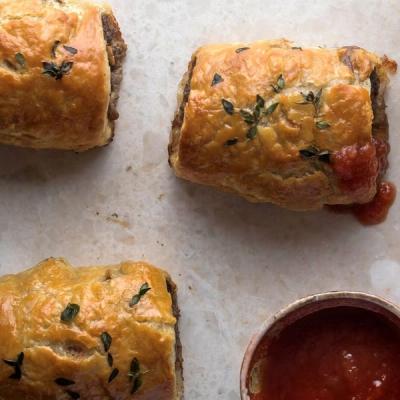 Three golden-brown sausage rolls garnished with herbs, served with red sauce. Three golden-brown sausage rolls garnished with herbs, served with red sauce.