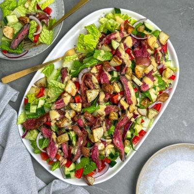 Steak salad with sliced steak, roasted potatoes, lettuce, cucumbers, capsicums, and red onions, drizzled with creamy dressing, served on a large white platter with a smaller bowl in the background.