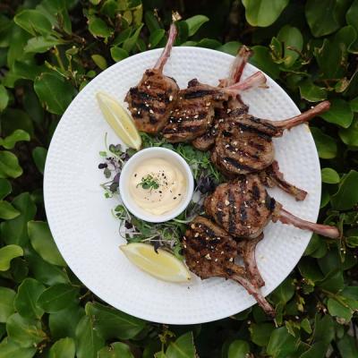 Grilled lamb chops with lemon, microgreens, and creamy sauce on a white plate, surrounded by green foliage. Grilled lamb chops with lemon, microgreens, and creamy sauce on a white plate, surrounded by green foliage.