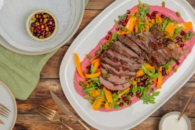 Sliced crusted lamb on a pink puree, garnished with rocket, carrot ribbons, pomegranate seeds, and pistachios on a white platter. Sliced crusted lamb on a pink puree, garnished with rocket, carrot ribbons, pomegranate seeds, and pistachios on a white platter.