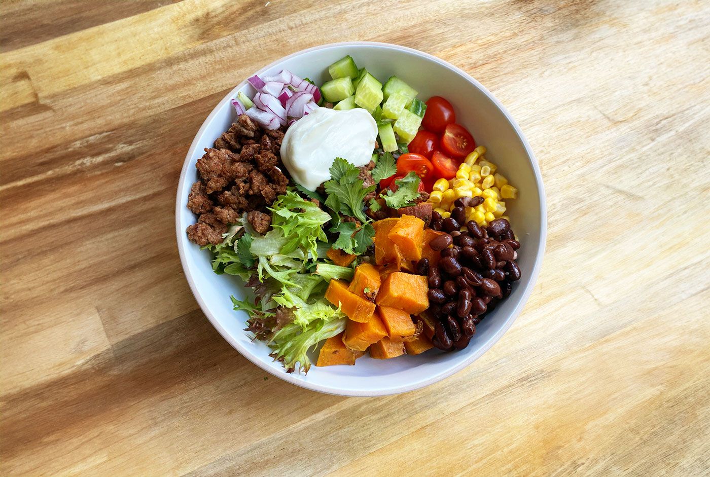 A vibrant burrito bowl with ground meat, sweet potatoes, black beans, corn, tomatoes, cucumber, and a dollop of sour cream on a wooden surface.