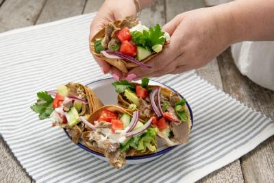 A person holds a gyro wrap filled with meat and fresh vegetables, with three more wraps on a plate below.