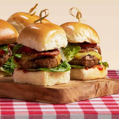 three lamb mini burgers are sitting on a wooden cutting board on a checkered table cloth. three lamb mini burgers are sitting on a wooden cutting board on a checkered table cloth.
