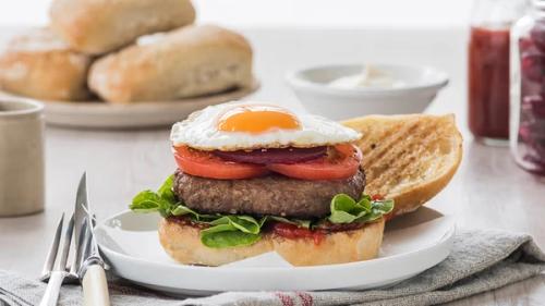 A burger topped with a fried egg, tomato, red onion, and lettuce, served open-faced with a toasted bun half on a white plate.