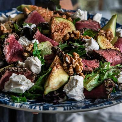 Salad with sliced rare beef, figs, walnuts, crumbled white cheese, and mixed greens on a blue patterned plate.