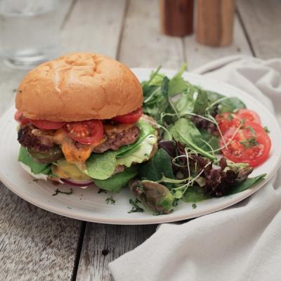 A lamb smash burger with tomato, lettuce, onion, and pickles, served with a side salad on a wooden table.