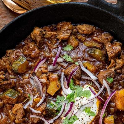 Close-up of a lamb curry in a cast iron pan, topped with red onion, cilantro, and sour cream. Close-up of a lamb curry in a cast iron pan, topped with red onion, cilantro, and sour cream.