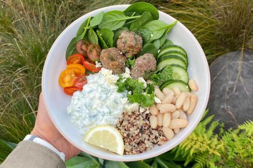 A vibrant, healthy bowl with meatballs, spinach, sliced cucumber, cherry tomatoes, white beans, quinoa, tzatziki, feta, and a lemon wedge, held outdoors.