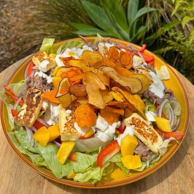 A colorful salad on a yellow plate, featuring lettuce, grilled halloumi cheese, lamb, mango chunks, red bell peppers, red onion, creamy white dressing, and a large pile of colorful kumara chips. A colorful salad on a yellow plate, featuring lettuce, grilled halloumi cheese, lamb, mango chunks, red bell peppers, red onion, creamy white dressing, and a large pile of colorful kumara chips.