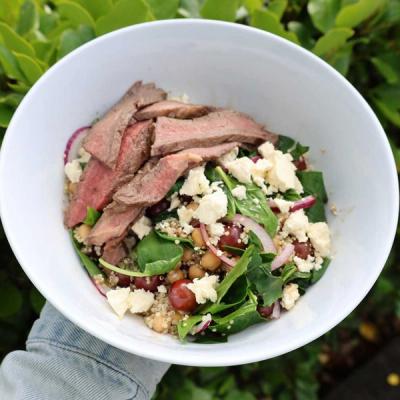 a person is holding a bowl of salad with steak , spinach, chickpeas, grapes and feta cheese.