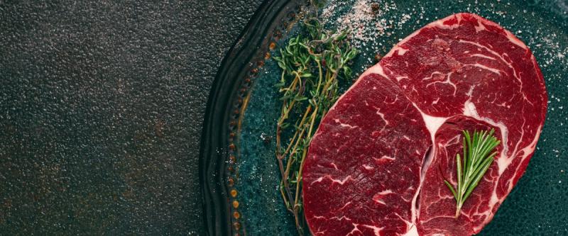 Raw steak with rosemary, thyme, and salt on a dark plate.