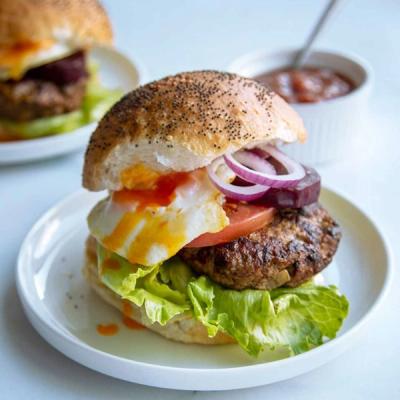 A burger with a poppy seed bun, grilled lamb patty, lettuce, tomato, red onion, and a fried egg with a runny yolk, served on a white plate.