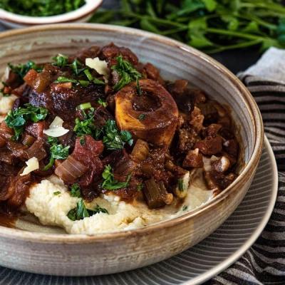 Bowl of braised osso buco over a creamy white base, garnished with herbs and cheese.