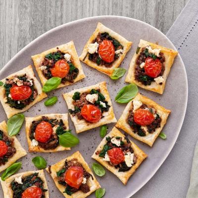 Puff pastry squares topped with ground meat, spinach, cherry tomatoes, and feta cheese, garnished with basil on a gray platter. Puff pastry squares topped with ground meat, spinach, cherry tomatoes, and feta cheese, garnished with basil on a gray platter.