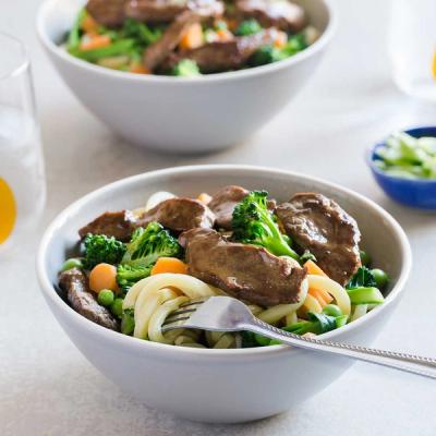 A bowl of udon noodles with lamb, broccoli, carrots, and peas, with a fork. A bowl of udon noodles with lamb, broccoli, carrots, and peas, with a fork.