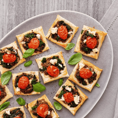 Square puff pastry appetizers topped with roasted tomatoes, spinach, and cheese, garnished with basil, on a grey platter. Square puff pastry appetizers topped with roasted tomatoes, spinach, and cheese, garnished with basil, on a grey platter.