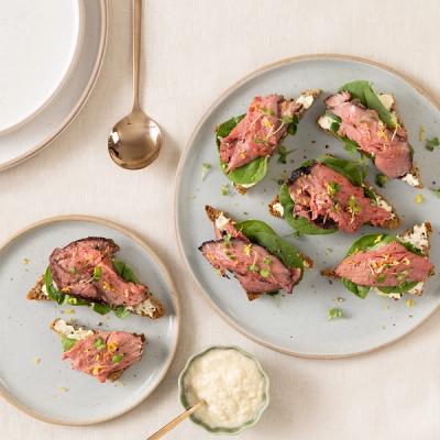 Open-faced roast beef sandwiches topped with spinach, horseradish, microgreens, and lemon zest on dark bread, served on grey plates.