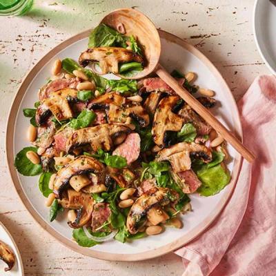 a white plate topped with a salad of steak, mushrooms, beans and spinach with a wooden spoon.
