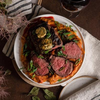 A seared steak, sliced medium-rare, served over mashed sweet potatoes, garnished with green onions and herb butter.