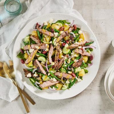 a white plate topped with a salad of lamb, green beans, cucumbers, olives and feta cheese.