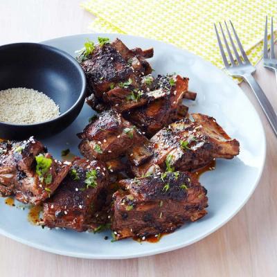 A light blue plate holds glazed lamb ribs topped with sesame seeds and herbs, with a small bowl of sesame seeds nearby.