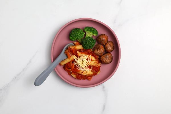 a pink plate topped with pasta , broccoli and meatballs with a spoon . a pink plate topped with pasta , broccoli and meatballs with a spoon .