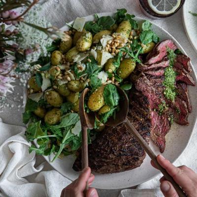 A plate of grilled steak with green sauce, and a potato salad with arugula, Parmesan, and pine nuts. Hands serve the salad with wooden spoons. A plate of grilled steak with green sauce, and a potato salad with arugula, Parmesan, and pine nuts. Hands serve the salad with wooden spoons.